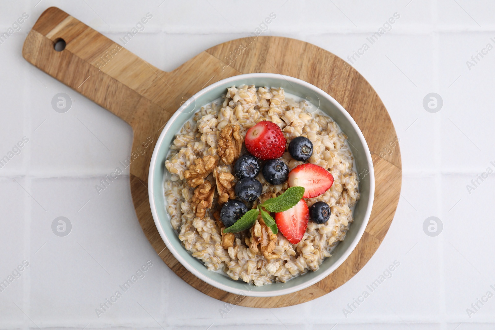 Photo of Tasty oatmeal with strawberries, blueberries and walnuts in bowl on white tiled table, top view