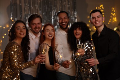 Photo of Happy friends with glasses of wine and sparklers celebrating birthday indoors, blurred view