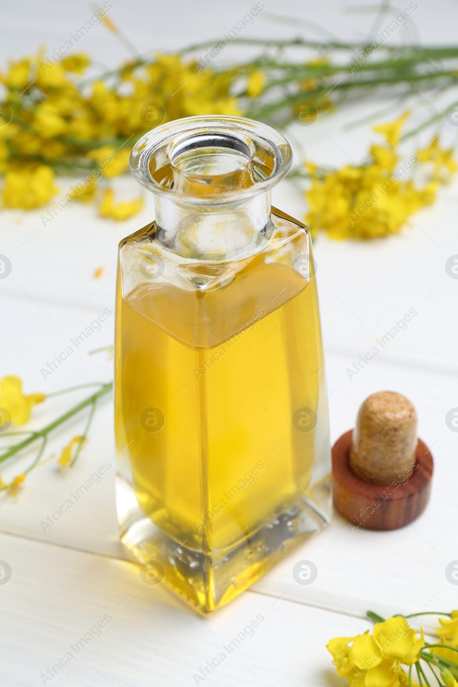 Photo of Rapeseed oil in glass bottle and beautiful yellow flowers on white wooden table, closeup