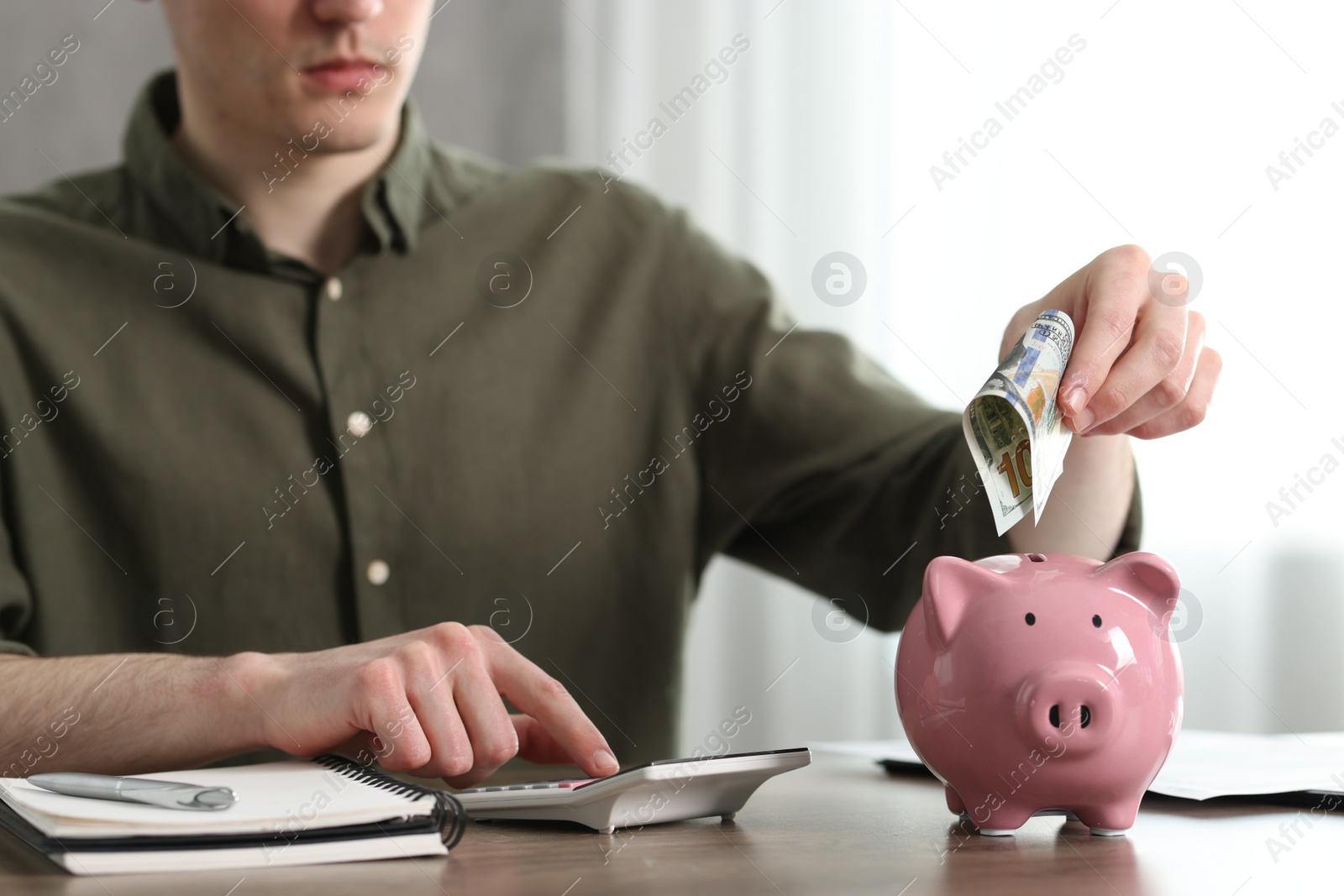 Photo of Financial savings. Man putting dollar banknote into piggy bank at wooden table, closeup