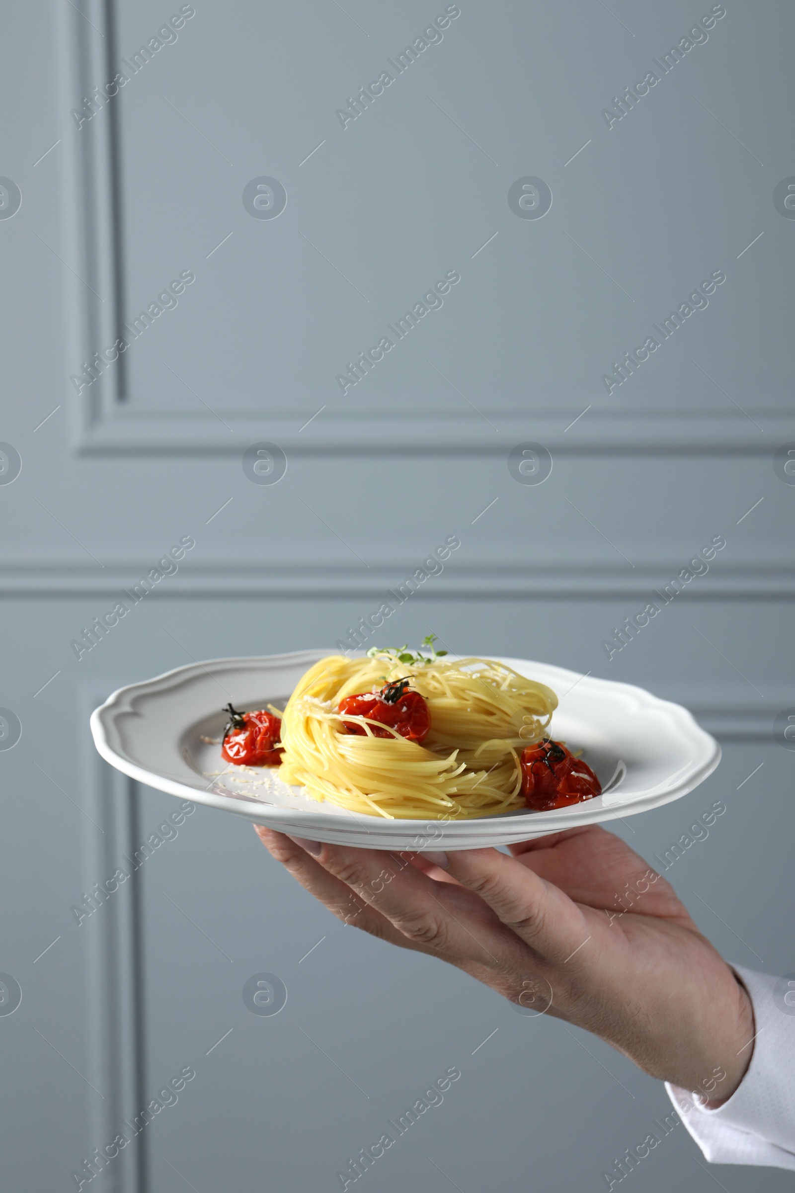 Photo of Waiter holding plate of tasty capellini with tomatoes and cheese near grey wall, closeup and space for text. Exquisite presentation of pasta dish