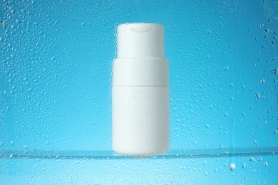 Photo of Bottle with moisturizing cream on light blue background, view through wet glass