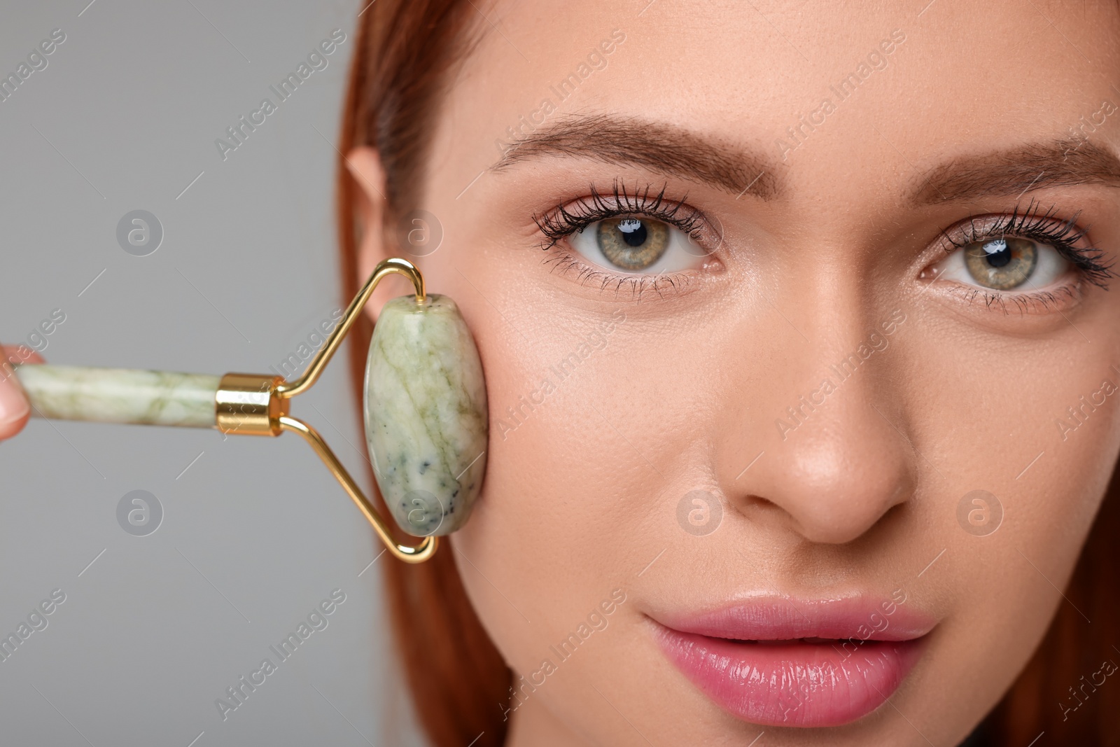 Photo of Young woman massaging her face with jade roller on grey background, closeup