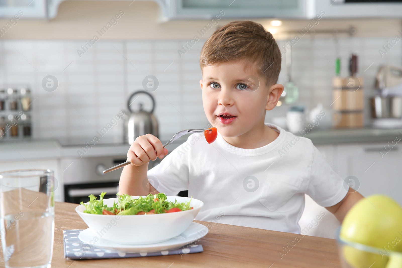 Photo of Adorable little boy eating vegetable salad at table in kitchen