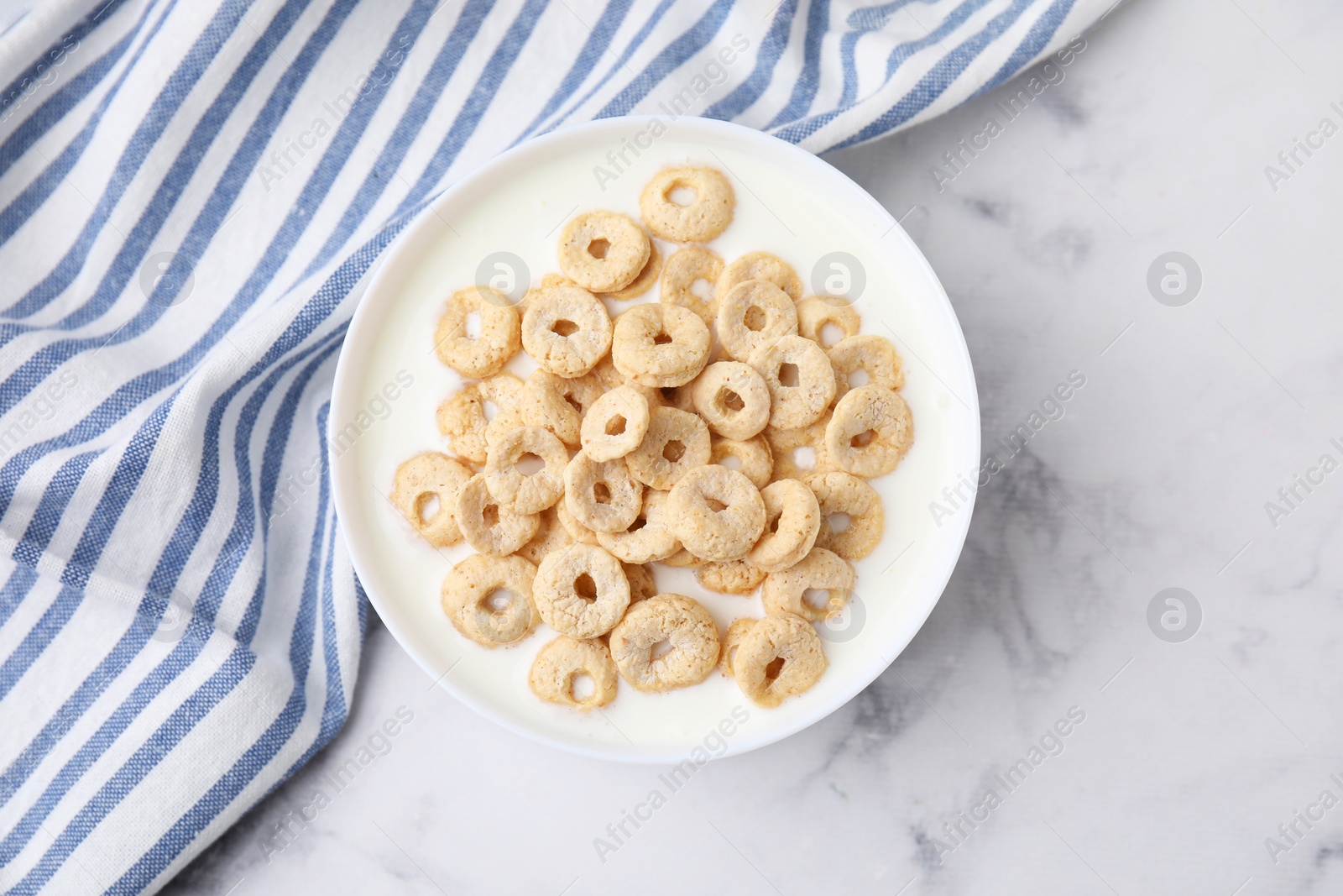 Photo of Breakfast cereal. Tasty corn rings with milk in bowl on white marble table, top view