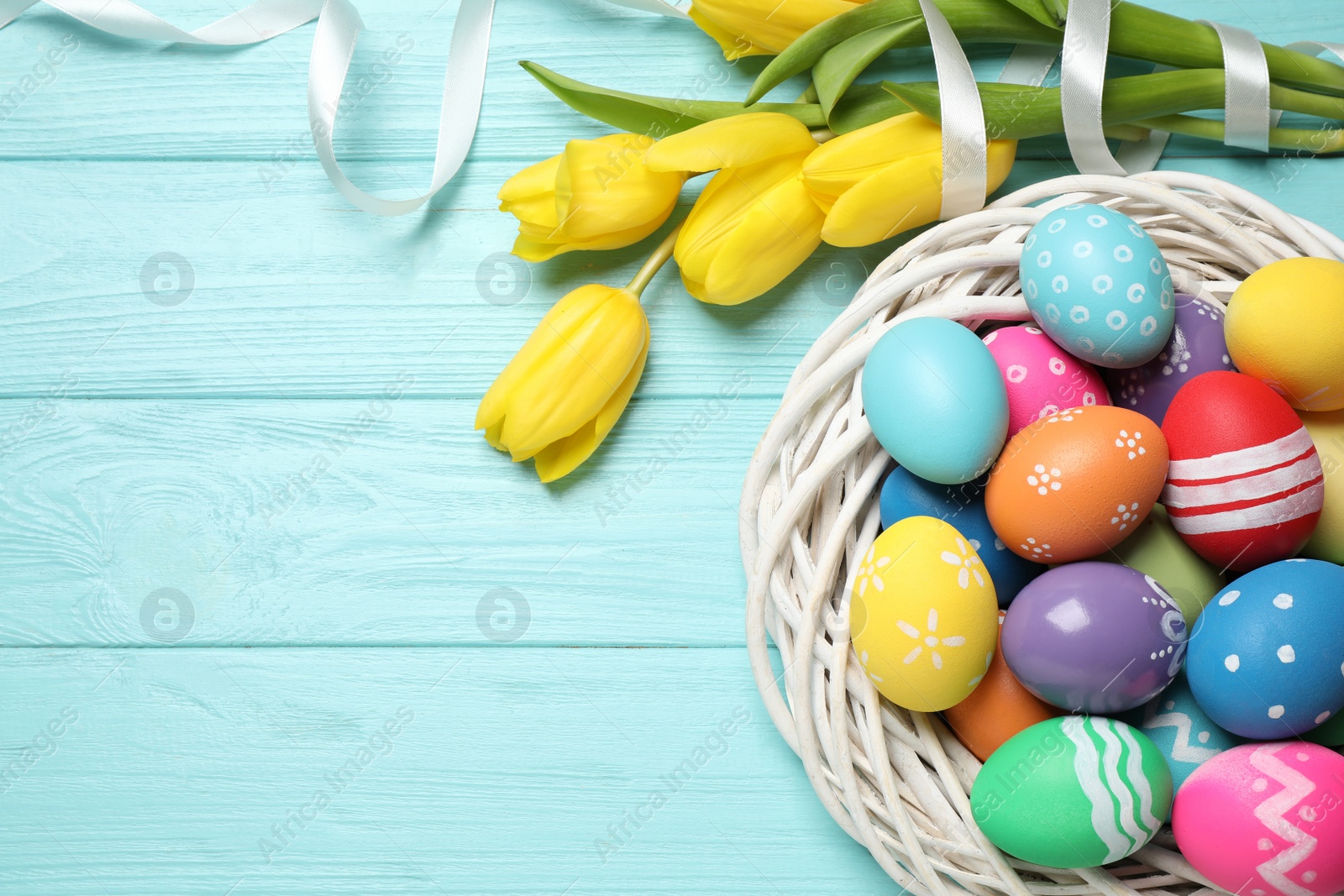 Photo of Colorful Easter eggs in decorative nest and tulip flowers on light blue wooden background, flat lay. Space for text