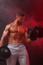 Photo of Young bodybuilder exercising with dumbbells in smoke on color background, low angle view