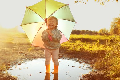 Little girl wearing rubber boots with colorful umbrella in puddle outdoors, space for text. Autumn walk