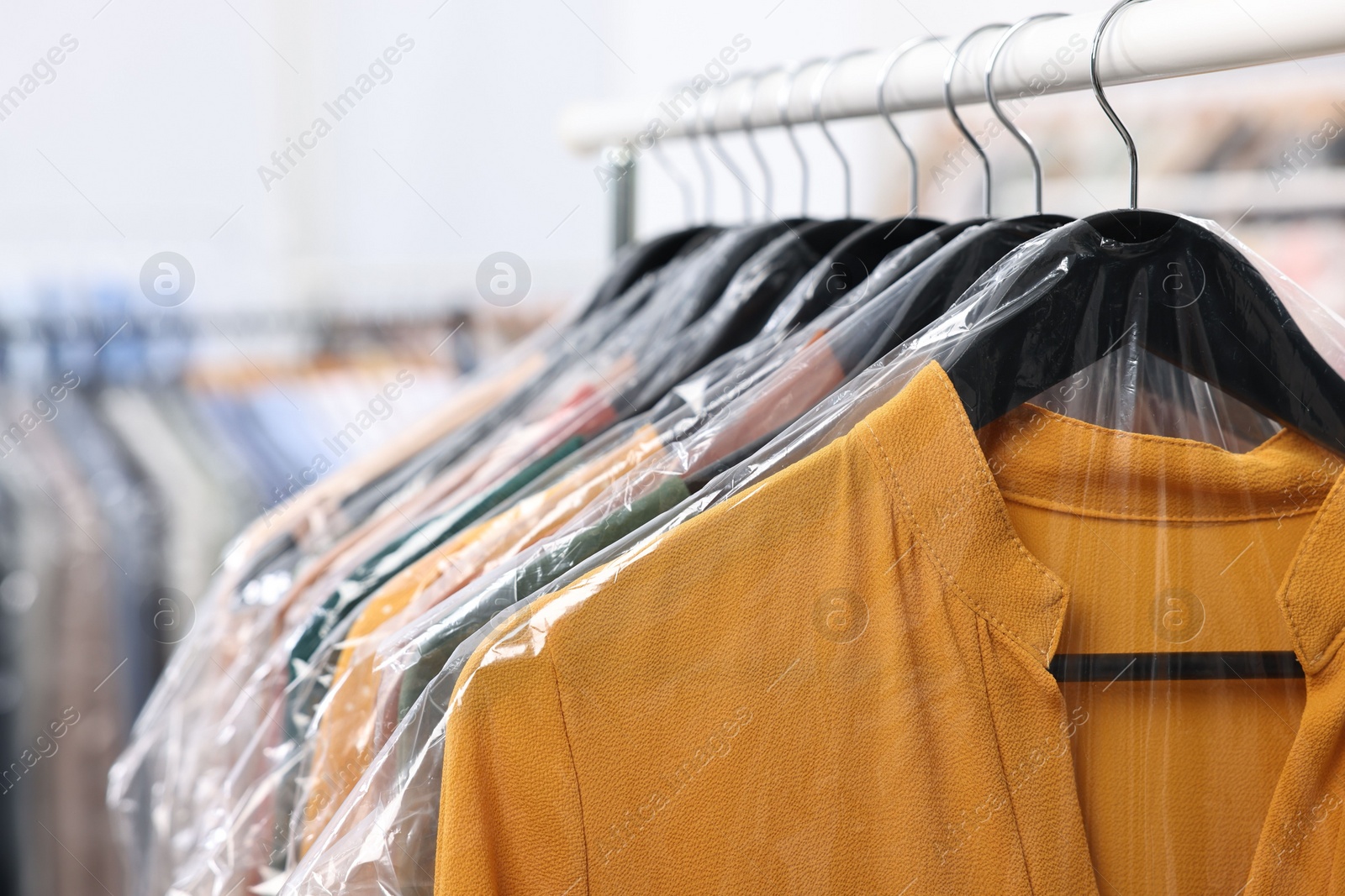 Photo of Dry-cleaning service. Many different clothes in plastic bags hanging on rack indoors, closeup