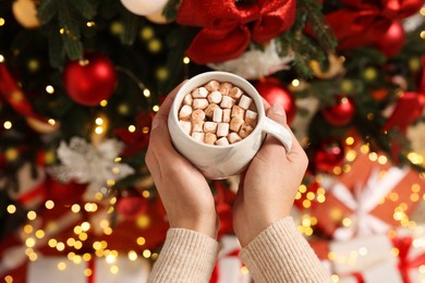 Woman holding cup of delicious cocoa with marshmallows near Christmas tree and gifts, above view
