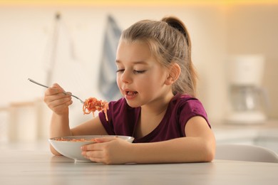 Cute little girl eating tasty pasta at table in kitchen