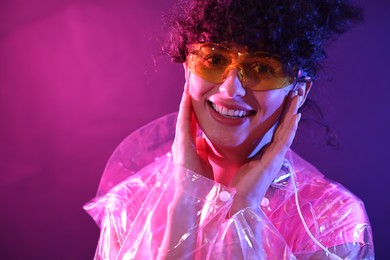 Photo of Beautiful young woman in transparent coat and sunglasses posing on color background in neon lights. Space for text