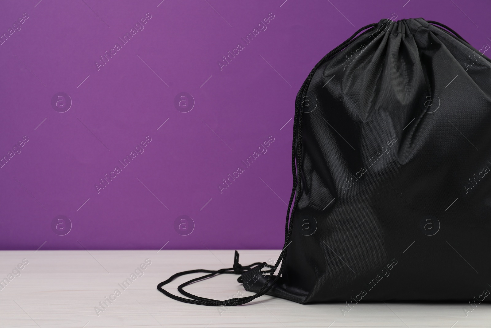 Photo of Black drawstring bag on white wooden table against purple background. Space for text