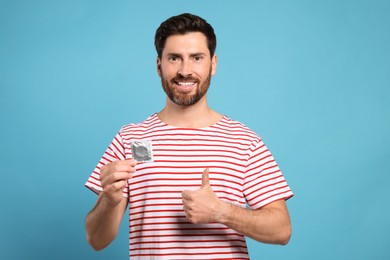 Happy man with condom showing thumb up on light blue background. Safe sex