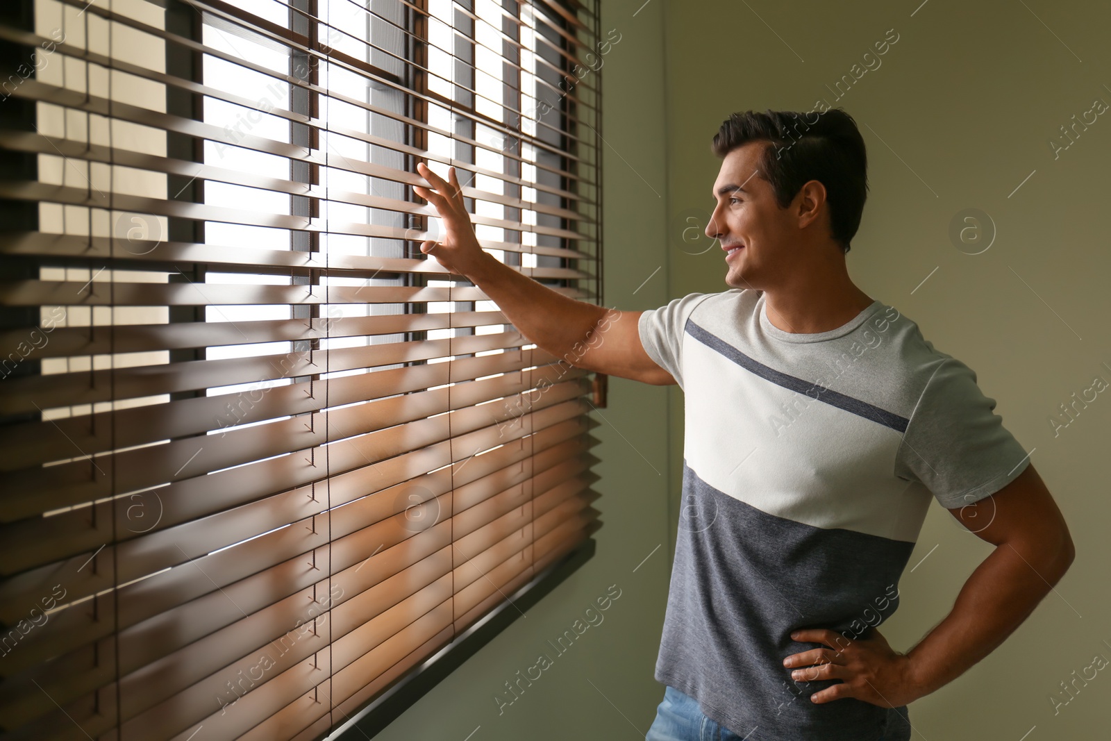 Photo of Handsome young man looking through window blinds indoors