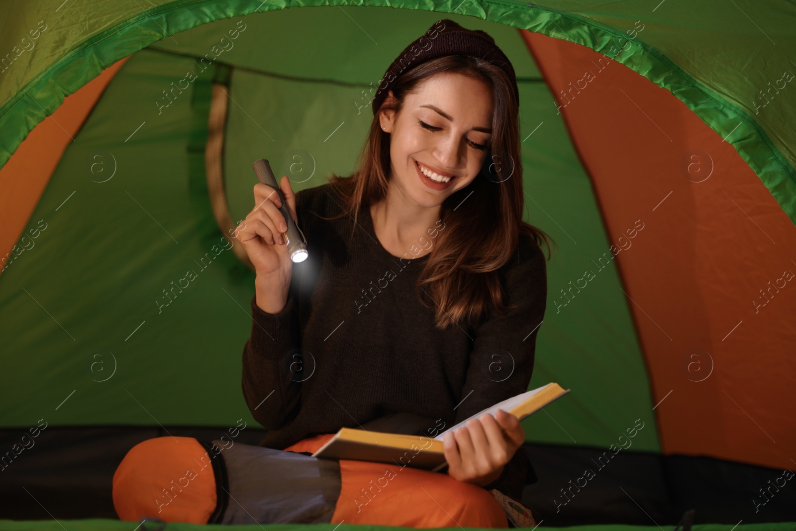 Photo of Young woman with flashlight reading book in tent