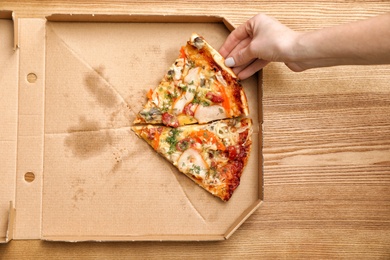 Woman taking pizza piece out of box on wooden table, top view