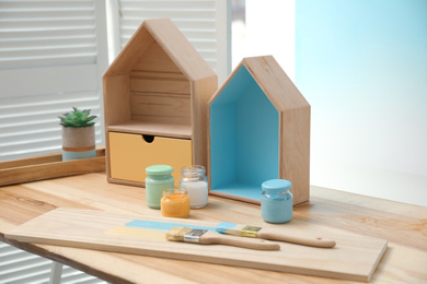 Photo of House shaped shelves, brushes and jars of paints on table indoors