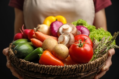 Photo of Woman holding wicker basket full of fresh vegetables on black background, closeup