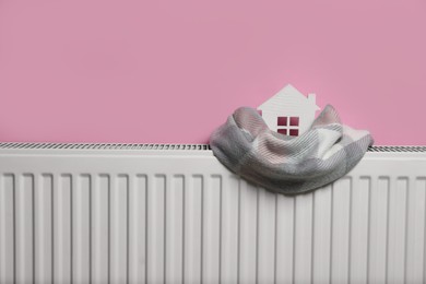 Photo of Modern radiator with knitted scarf and wooden house near pink wall indoors, space for text. Winter heating efficiency