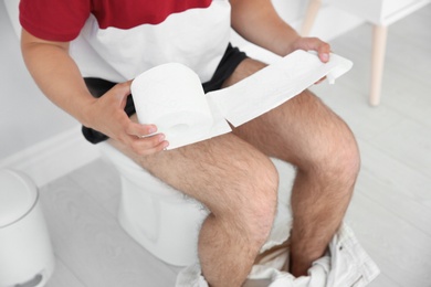 Photo of Young man with bath tissue sitting on toilet bowl at home