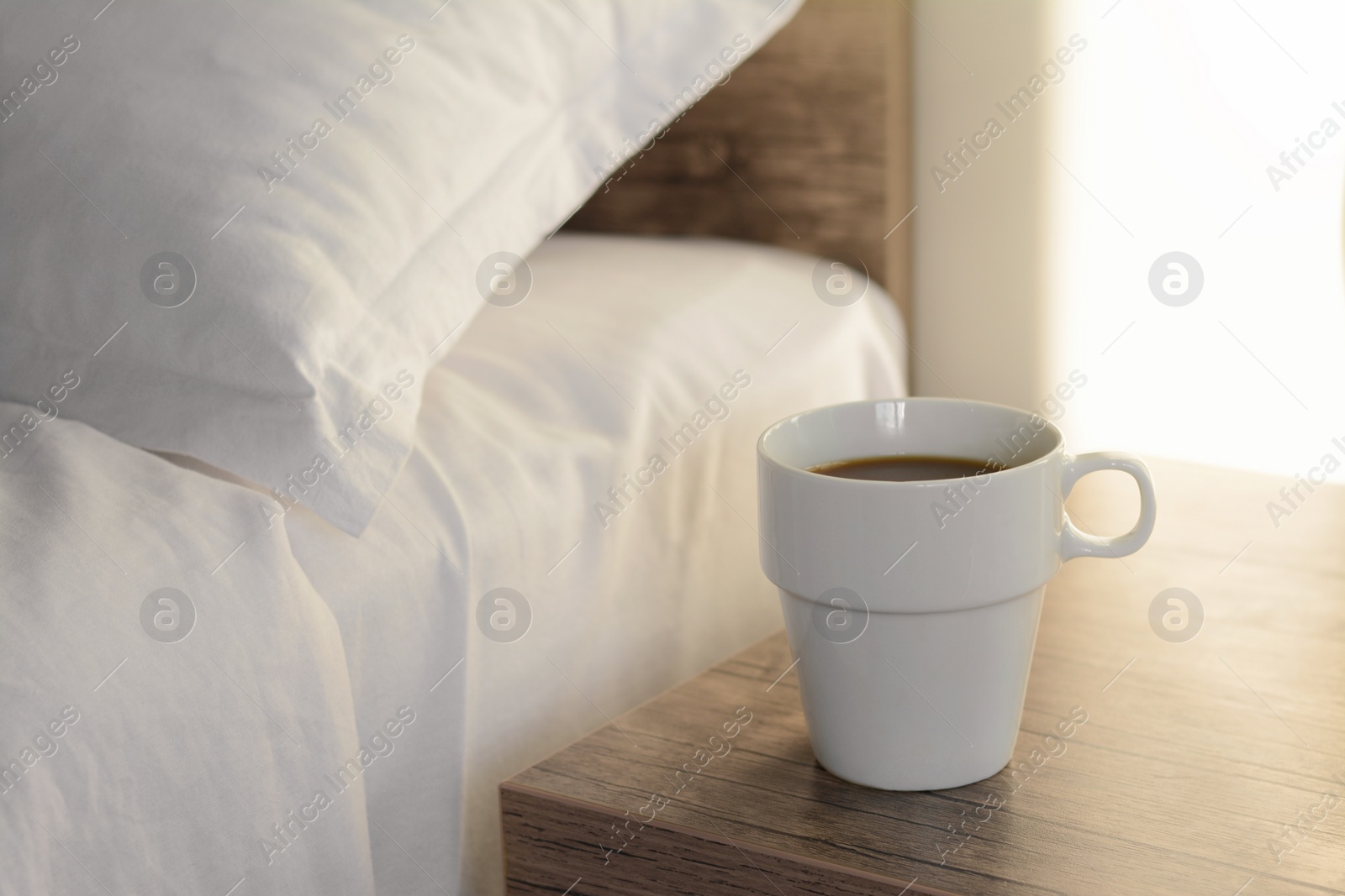 Photo of Cup of morning coffee on wooden night stand near bed indoors