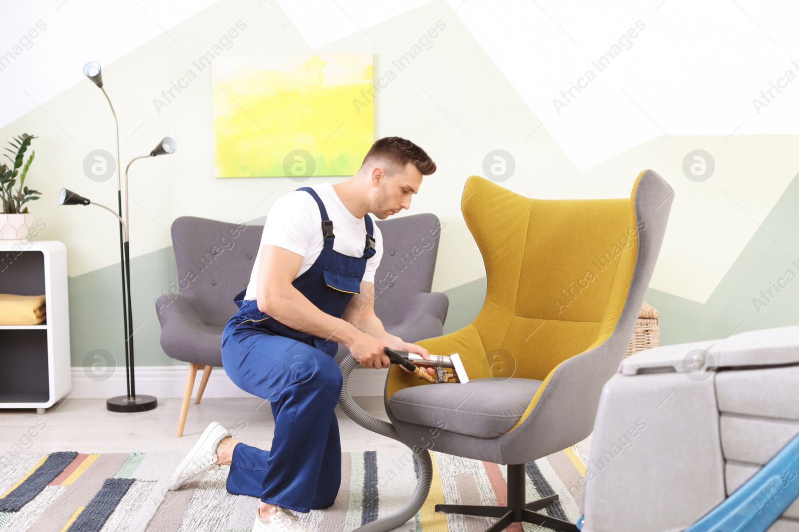 Photo of Male worker removing dirt from armchair with professional vacuum cleaner indoors