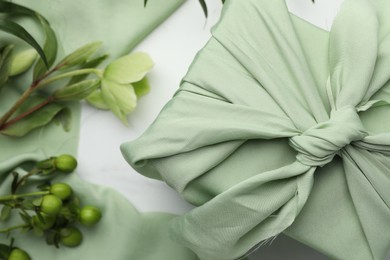 Photo of Furoshiki technique. Gift packed in green fabric and plants for decor on white table, flat lay