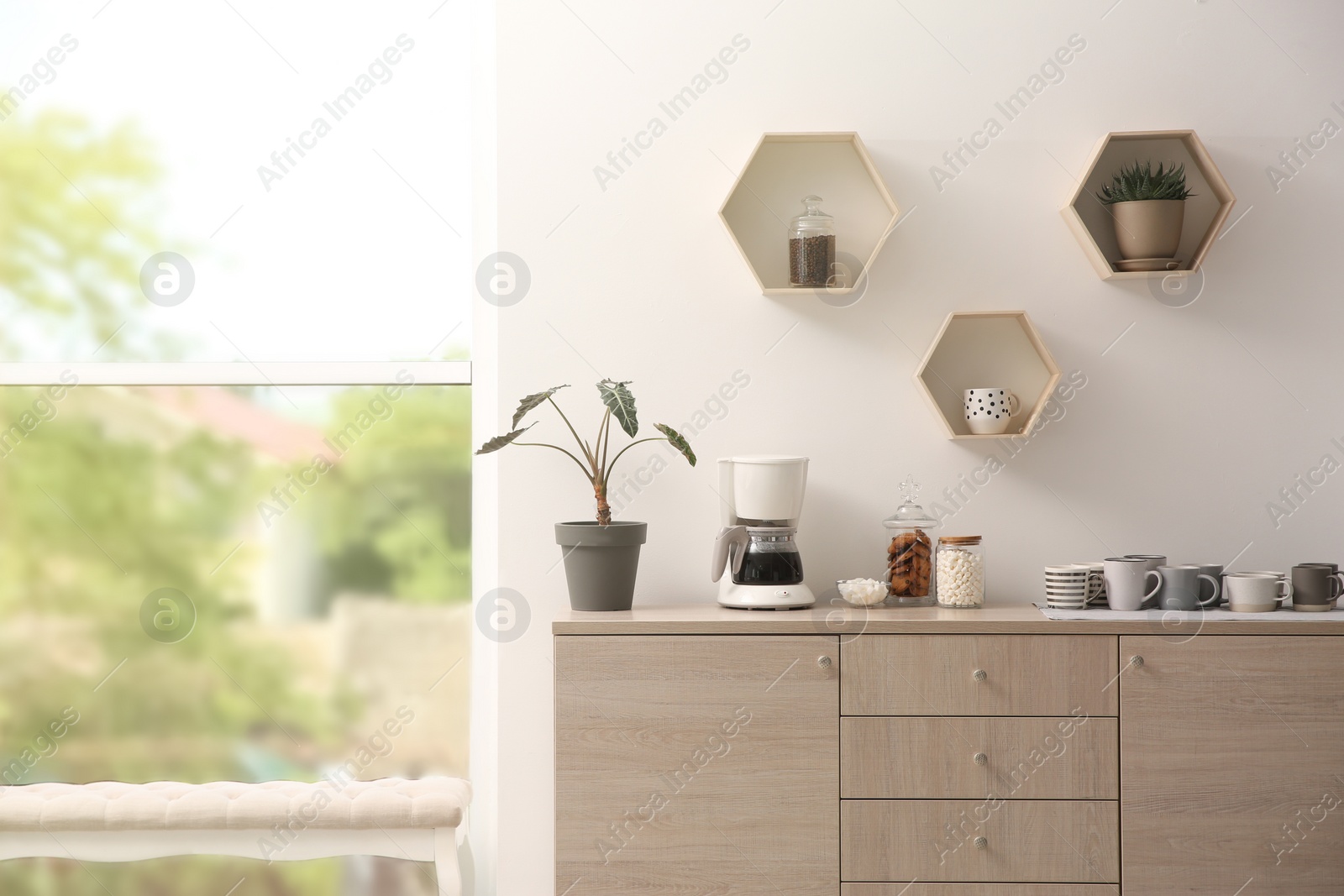 Photo of Modern coffeemaker and cups on commode near light wall indoors