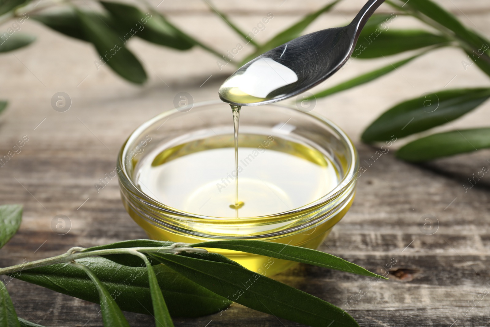 Photo of Pouring olive oil from spoon into glass bowl on wooden table, closeup