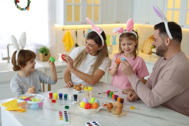 Photo of Easter celebration. Happy family with bunny ears having fun while painting eggs at white marble table in kitchen