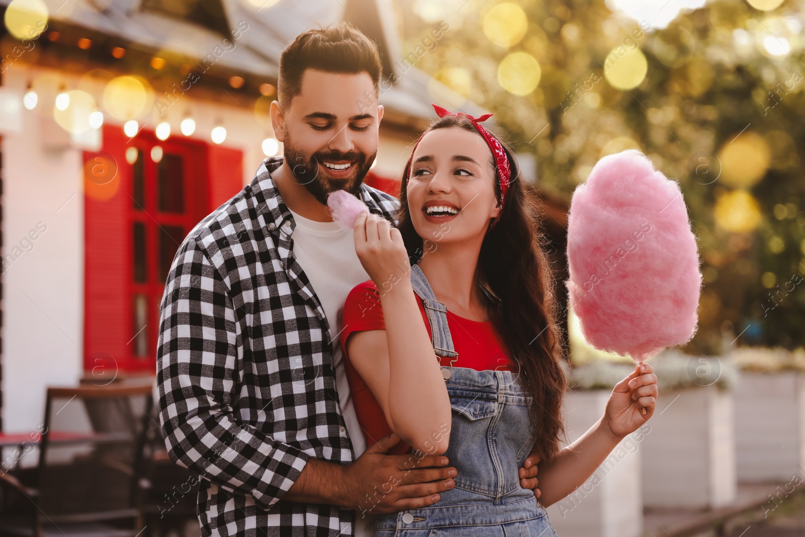 Image of Happy young woman treating her boyfriend with cotton candy outdoors on sunny day