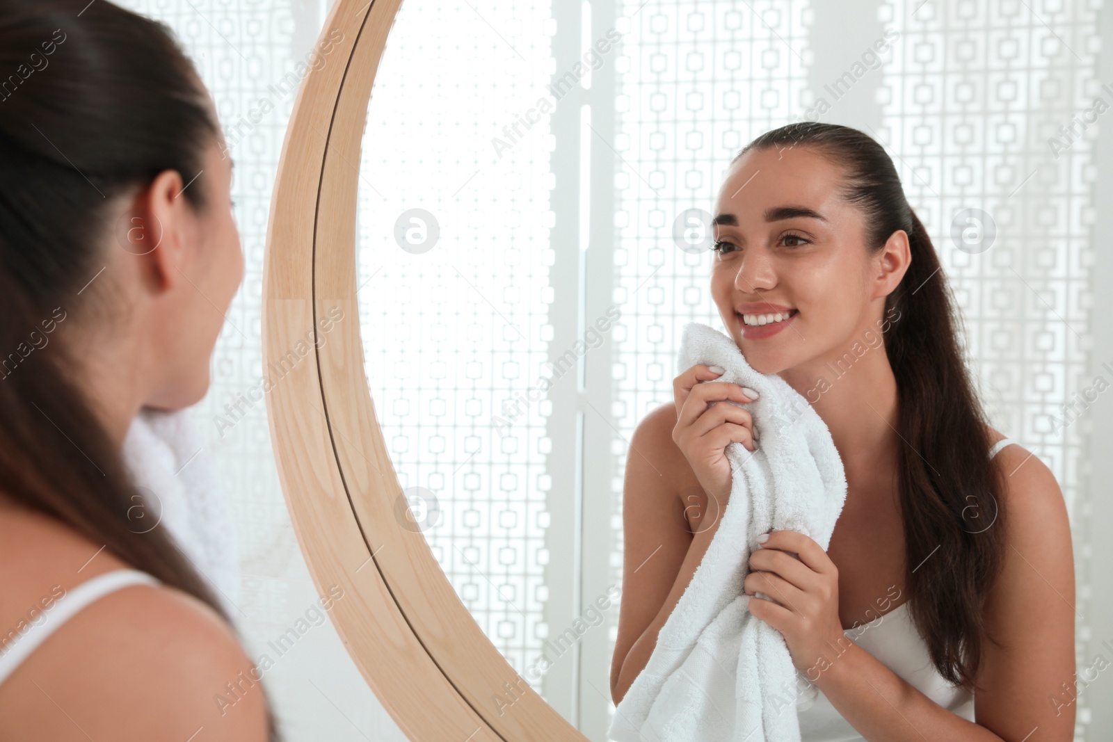 Photo of Beautiful young woman wiping face with towel near mirror in bathroom. Facial wash