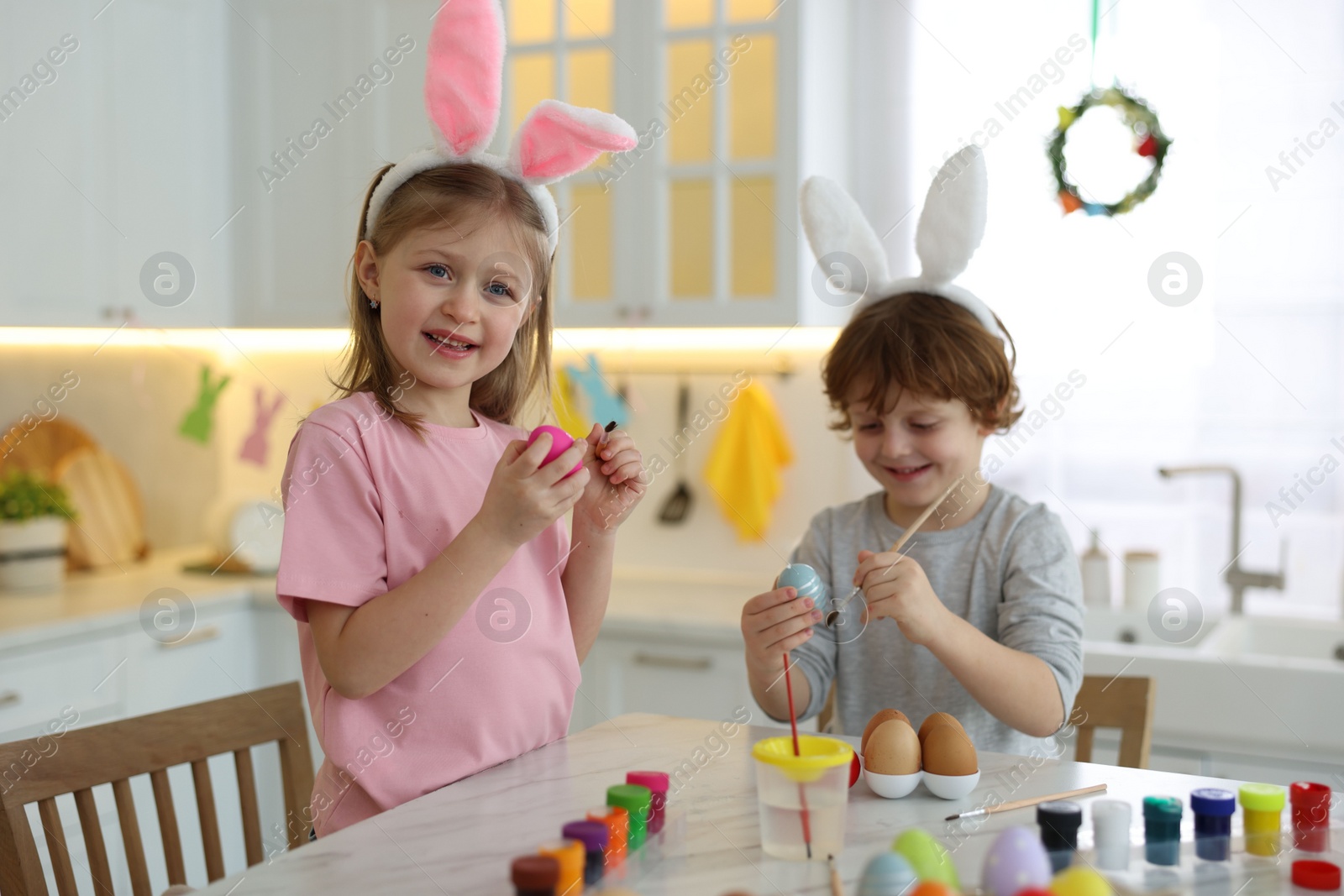 Photo of Easter celebration. Cute children with bunny ears painting eggs at white marble table in kitchen