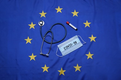 Stethoscope, medical mask with word COVID-19 and test tube on flag of European Union, flat lay