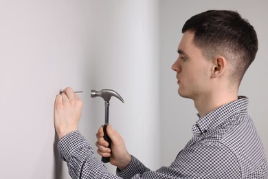 Young man hammering nail into white wall indoors