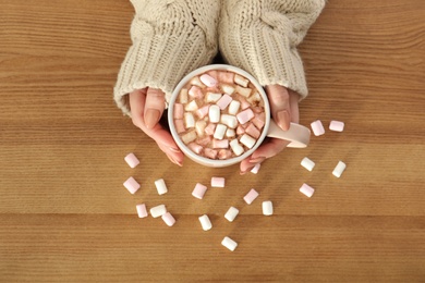 Photo of Woman holding cup of aromatic cacao with marshmallows on wooden background, top view