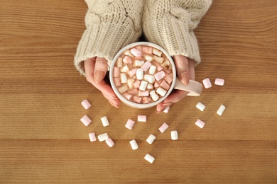 Woman holding cup of aromatic cacao with marshmallows on wooden background, top view