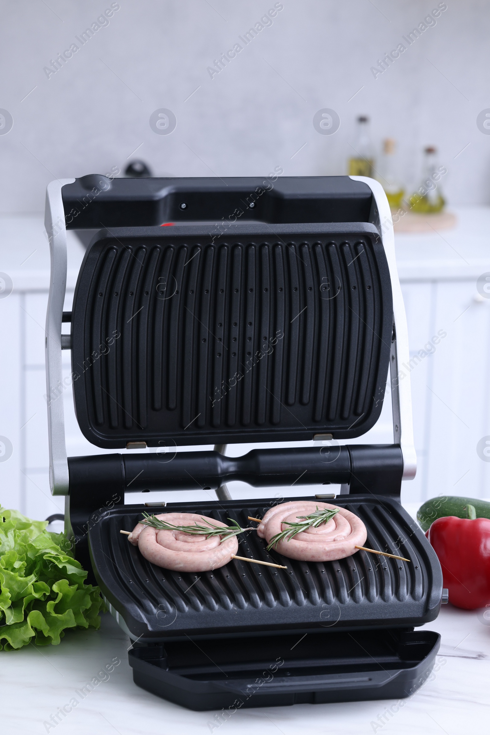 Photo of Electric grill with homemade sausages, rosemary and vegetables on white table in kitchen