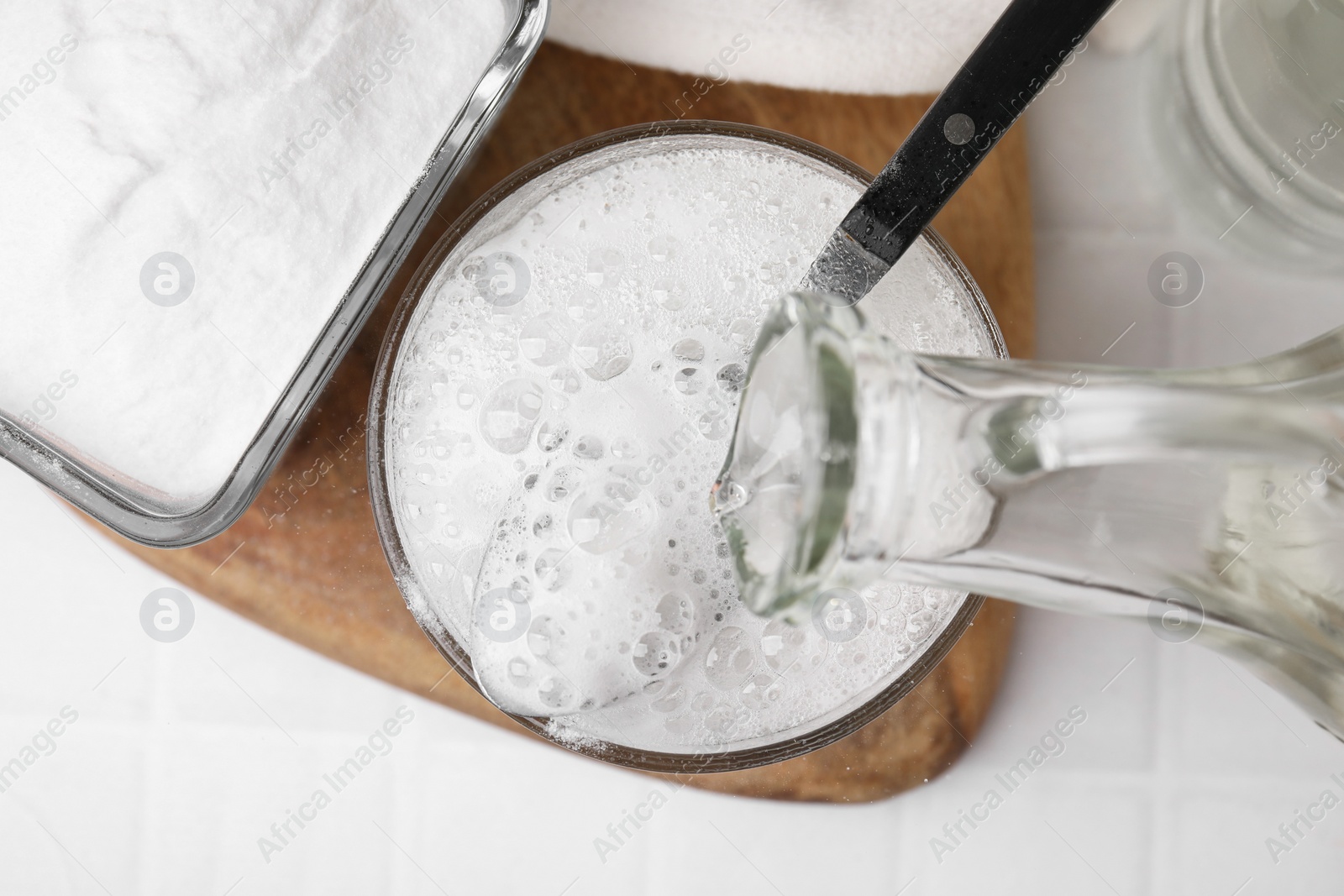 Photo of Pouring vinegar into spoon with baking soda over bowl at white tiled table, top view