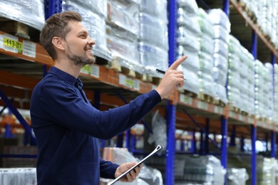 Photo of Happy manager holding clipboard in warehouse with lots of products. Space for text