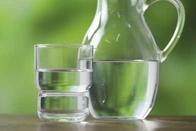 Photo of Jug and glasses with clear water on wooden table against blurred green background, closeup