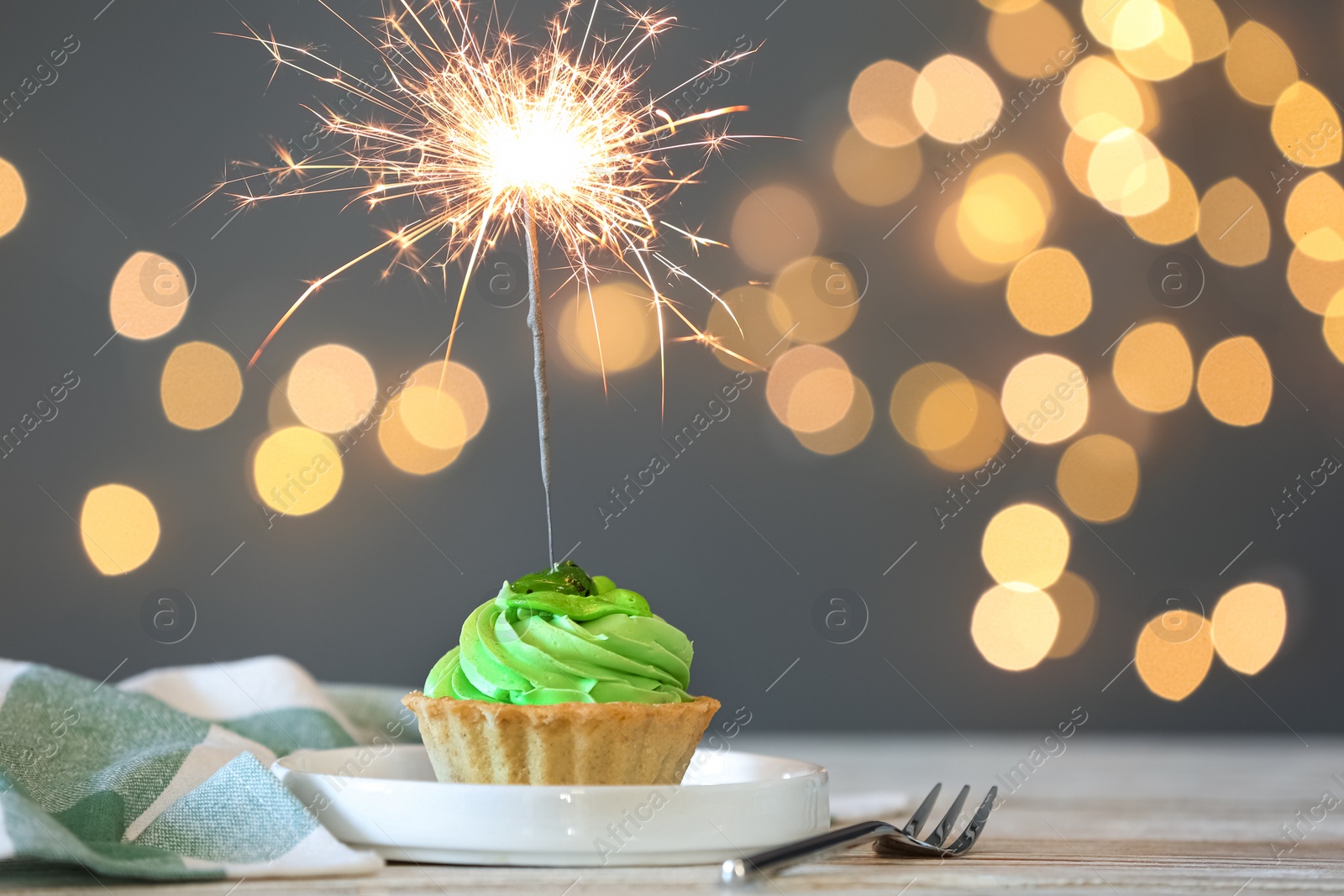 Photo of Cupcake with burning sparkler on table against blurred festive lights. Space for text