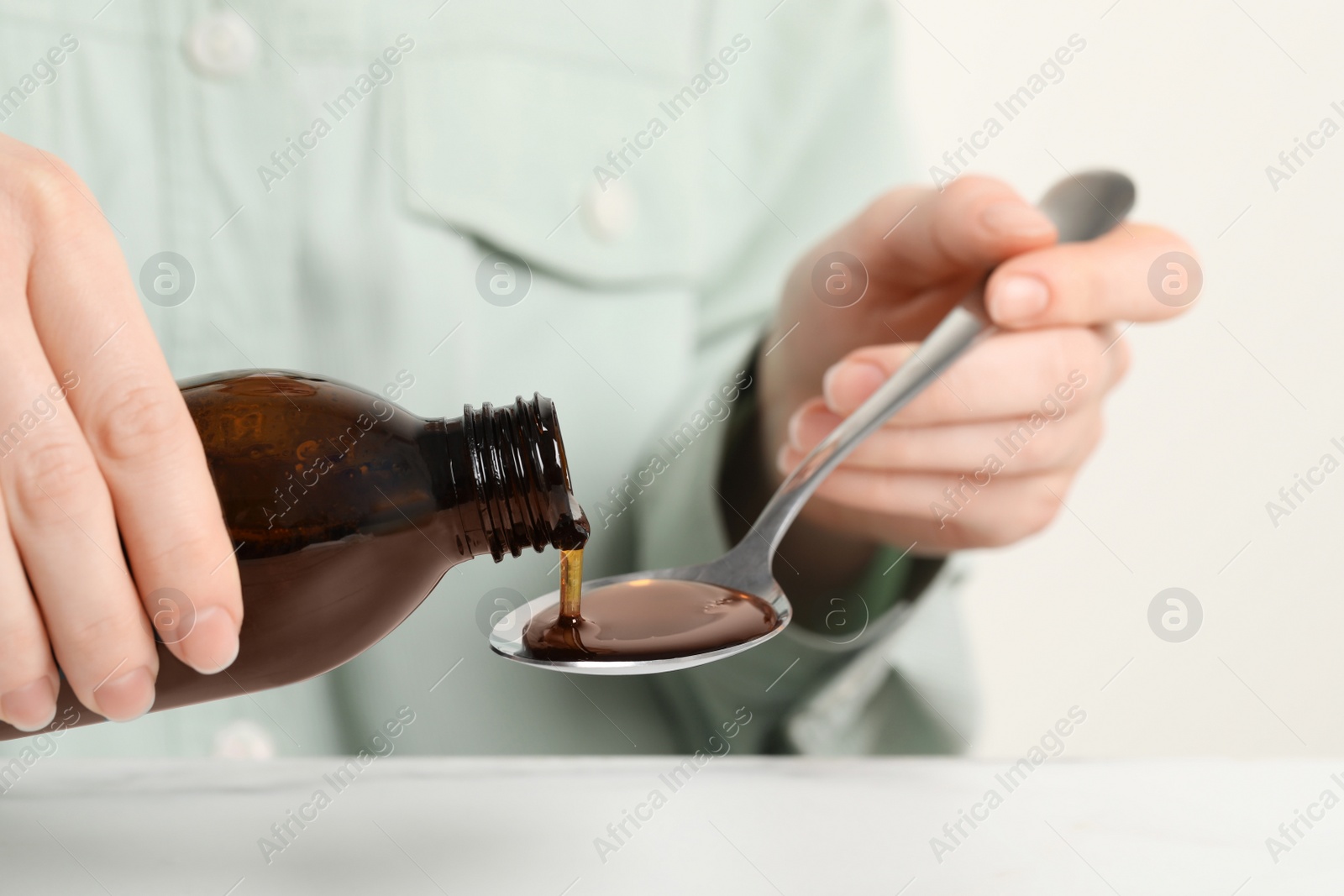 Photo of Woman pouring syrup from bottle into spoon at table, closeup. Cold medicine