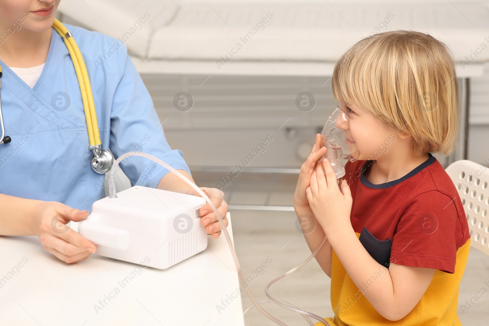 Photo of Medical assistant sitting near sick little boy while he using nebulizer for inhalation in hospital