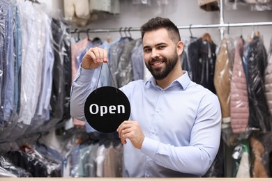 Photo of Dry-cleaning service. Happy worker holding Open sign at counter indoors