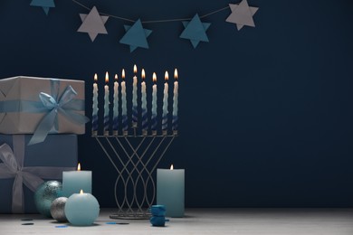 Photo of Hanukkah celebration. Menorah, burning candles, dreidels and gift boxes on white table against blue background, space for text