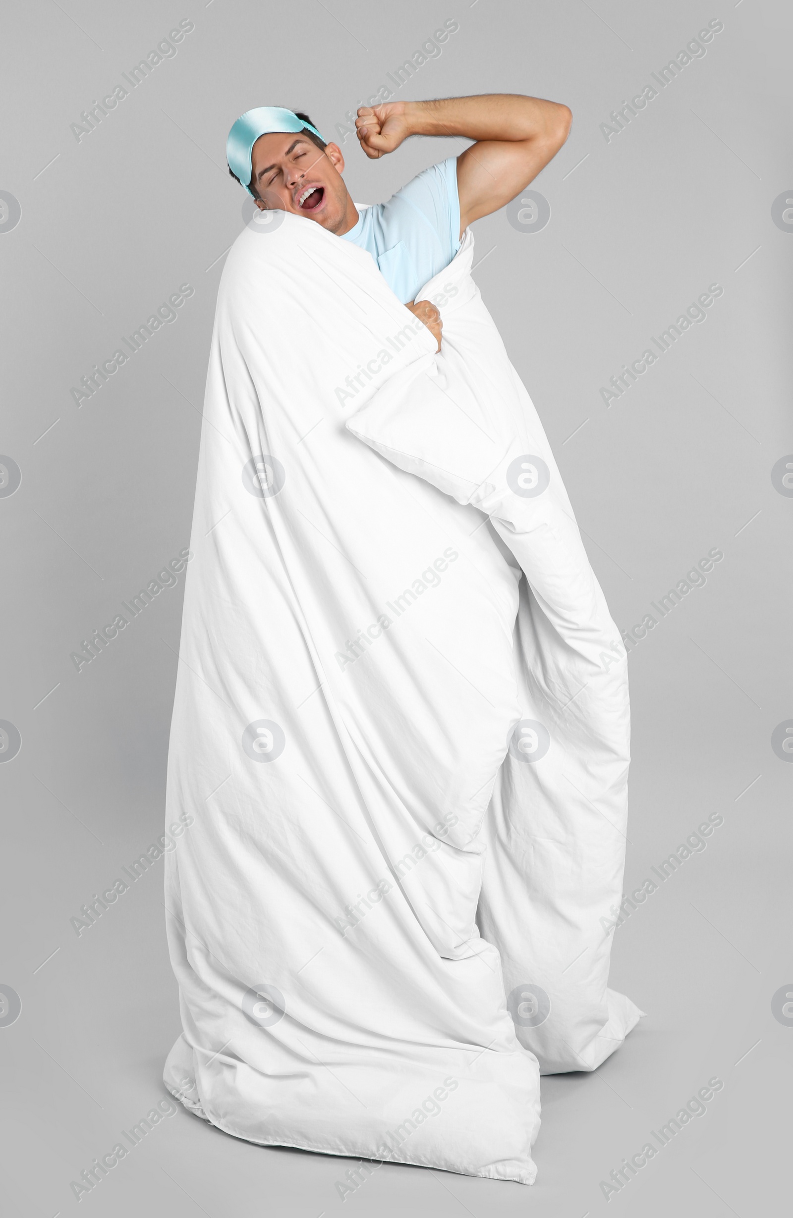 Photo of Man in sleeping mask wrapped with blanket yawning on grey background