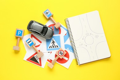 Photo of Many different road signs, notebook with sketch of roundabout and toy car on yellow background, flat lay. Driving school