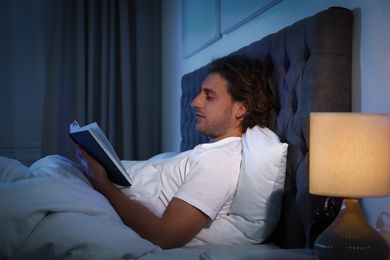 Photo of Handsome young man reading book in dark room at night. Bedtime
